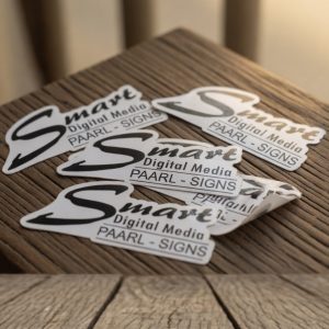 Collection of die-cut vinyl stickers featuring the Smart Digital Media PAARL - SIGNS logo arranged on a dark wooden table, with one sticker partially peeled and curling upward at the corner.